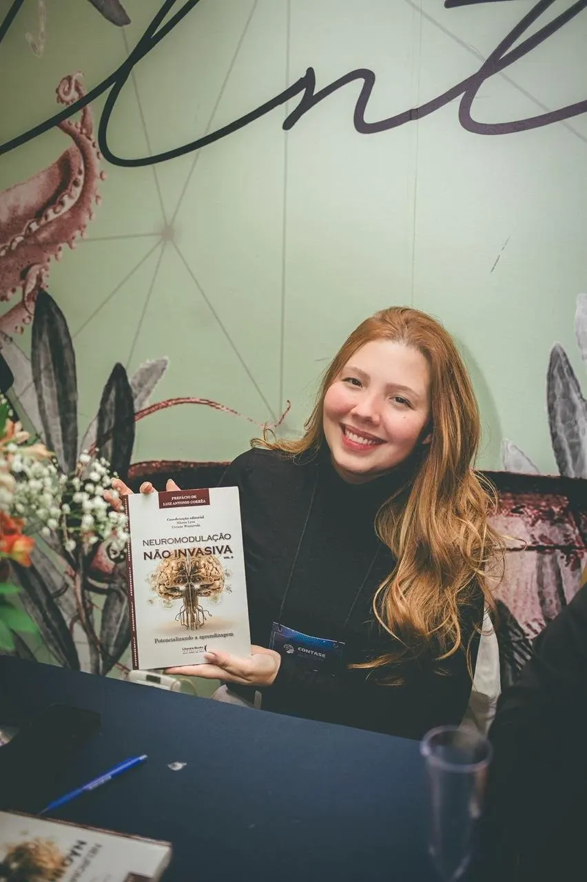 Brunna Vasconcellos sorrindo com o livro Neuromodulação Não Invasiva vol. 2, no Congresso Contase